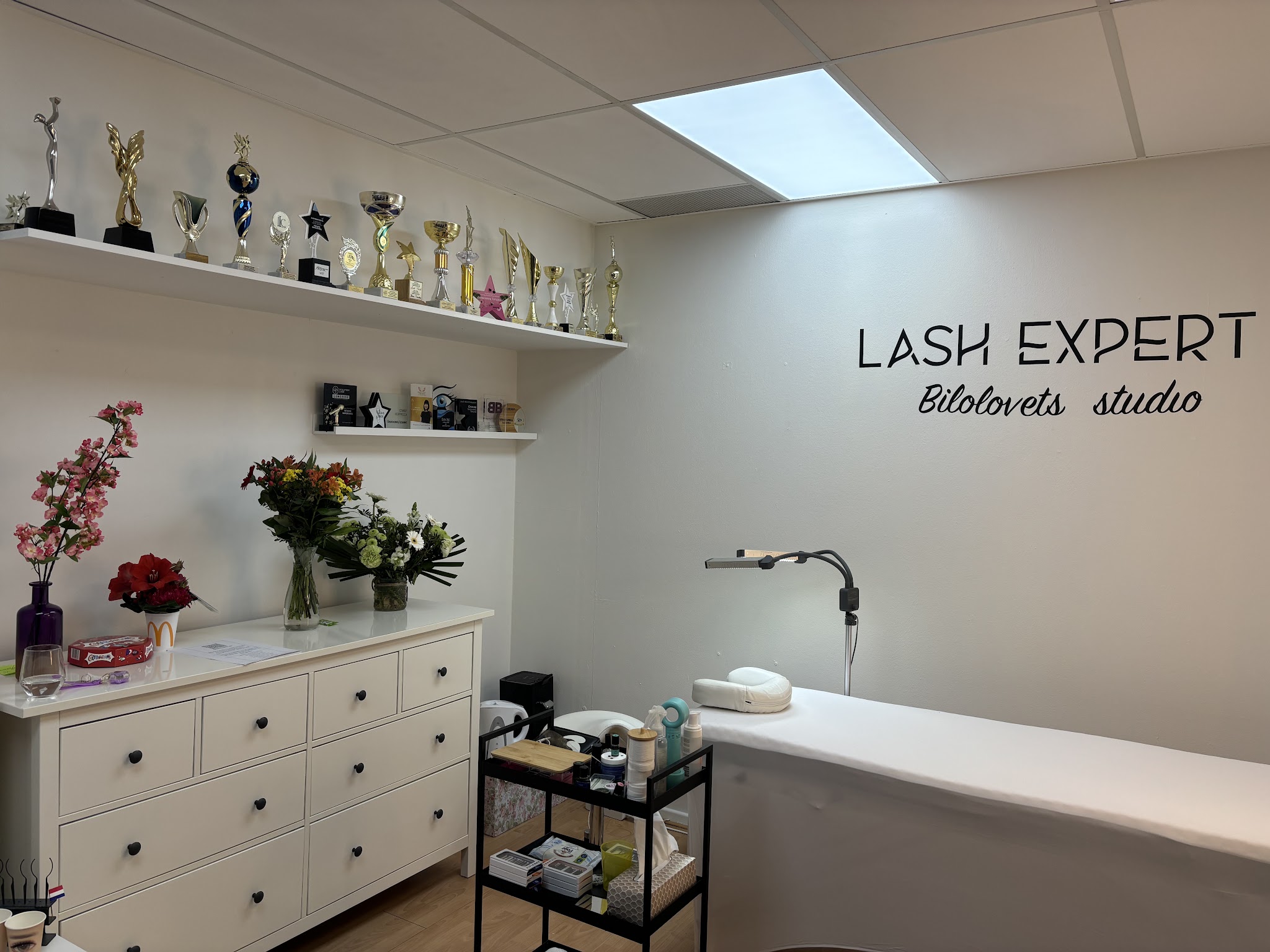De studio met de behandelbank, witte ladekasten, bloemen en een plank vol bekers onder de letters LASH EXPERT Bilolovets studio